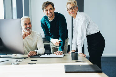 Drei fröhlich lächelnde Mitarbeiter arbeiten zusammen und tauschen Geschäftsideen für ihr Unternehmen aus. Eine Gruppe von Geschäftsleuten arbeitet in einem modernen Büro um einen Computer herum. - JLPPF01897