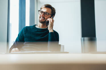 Fröhlicher junger Geschäftsmann sitzt an seinem Schreibtisch und spricht mit einem Mobiltelefon, er trägt elegante Businesskleidung und eine Brille. Professioneller Geschäftsmann kommuniziert mit seinen Arbeitskollegen. - JLPPF01899