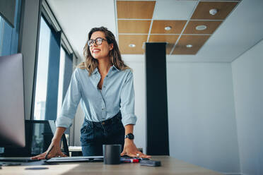 Eine Geschäftsfrau blickt nachdenklich an ihrem Schreibtisch in einem professionellen Büro vorbei. Sie trägt ihre elegante Geschäftskleidung und ist bereit, die Verantwortung zu übernehmen. - JLPPF01945