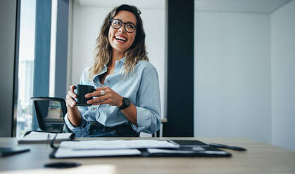 Young business woman and entrepreneur sits at her desk, smiling happily while holding a cup of coffee. Professional woman wearing eyeglasses and smart businesswear in an office. - JLPPF01946