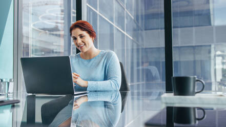 Female business executive conducts a virtual meeting using her laptop, confidently discussing important business matters with her team. Business woman sitting at a boardroom table in an office. - JLPPF01959