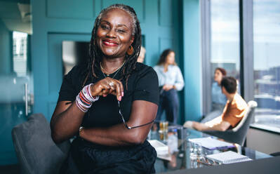 Confident black woman standing in a modern office space during a productive business meeting. Mature female professional smiling at the camera with her colleagues discussing in the background. - JLPPF01975