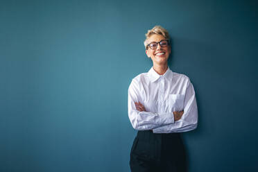 Successful finance professional looking at the camera with pride and happiness in an office. Mature business woman standing against a blue wall with crossed arms, wearing eyeglasses and businesswear. - JLPPF01980