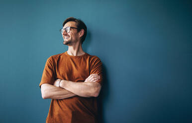 Erfolgreicher Geschäftsmann lächelt, während er vor einer blauen Wand in einem Büro steht. Männlicher Geschäftsmann, der über seine Leistungen in seinem Unternehmen nachdenkt und sich darauf freut, noch mehr zu erreichen. - JLPPF01989