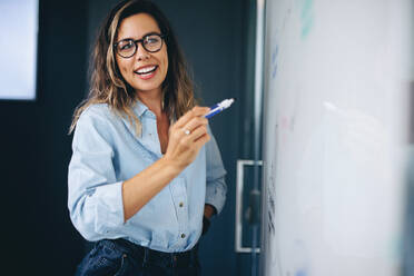 Eine fröhliche Geschäftsfrau steht vor einem Whiteboard und präsentiert begeistert ihre Ideen während einer Teambesprechung. Eine junge Berufstätige teilt ihre Erkenntnisse und Visionen mit ihren Kollegen. - JLPPF02000