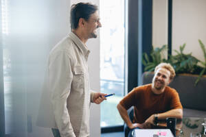 Professioneller Mann, der glücklich lächelt, während er vor seinem Team steht und seine Geschäftsstrategie präsentiert. Erfolgreicher Geschäftsmann, der sich mit seinen Kollegen unterhält, während sie ein Projekt in einem Büro planen. - JLPPF02011
