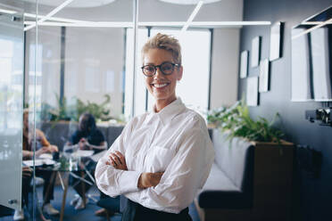 Glückliche Unternehmerin, die in die Kamera lächelt, während sie vor einem Besprechungsraum in einem modernen Büro steht. Reife Geschäftsfrau, die an einer Konferenz teilnimmt und sich mit ihren Geschäftspartnern trifft. - JLPPF02017