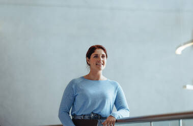 Young business woman with red hair looking away as she stands on an interior balcony in a modern workplace. Cheerful female professional thinking of her next business move. - JLPPF02025