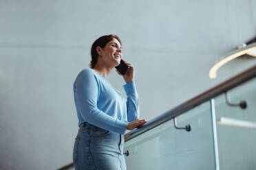 Eine Geschäftsfrau lächelt, als sie auf einem Balkon in einem Büro steht und mit ihren Kunden telefoniert. Eine junge Berufstätige, die erfolgreiche Geschäftsbeziehungen für ihr Unternehmen knüpft. - JLPPF02027