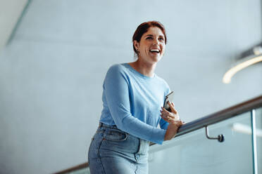 Erfolg und Selbstbestimmung von Frauen in der Unternehmenswelt: Eine junge Geschäftsfrau und Unternehmerin steht mit einem glücklichen Lächeln auf dem Balkon eines Büros und hält ein Mobiltelefon in der Hand. - JLPPF02029
