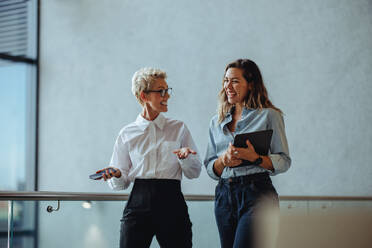 Successful female executives discuss their goals and plans as they walk together in an office. Two business women smile happily while working together in a professional setting. - JLPPF02038