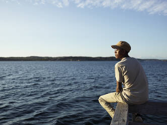 Junger Mann sitzt auf Holzreling am Meer - FOLF12227