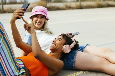 Happy woman sharing smart phone with friend sitting on footpath - EBSF03313