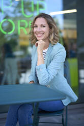 Lächelnde Geschäftsfrau mit Hand am Kinn, die in einem Straßencafé sitzt - PNEF02826