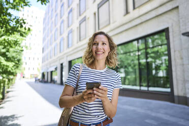 Smiling woman holding smart phone walking near building - PNEF02829