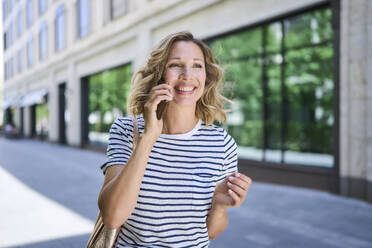 Glückliche blonde Frau, die in der Nähe eines Gebäudes mit einem Mobiltelefon spricht - PNEF02831