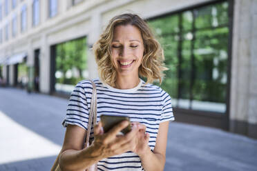 Glückliche Frau mit blondem Haar, die ein Smartphone in der Nähe eines Gebäudes benutzt - PNEF02832
