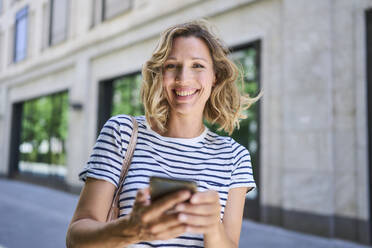 Happy woman with blond hair holding smart phone - PNEF02833