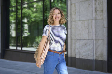 Smiling woman with bag walking near building - PNEF02835