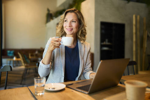 Smiling businesswoman having coffee sitting with laptop at cafe - PNEF02838