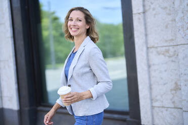 Happy businesswoman holding disposable cup in front of building - PNEF02843