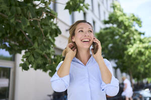 Glückliche Frau mit Hand im Haar, die mit einem Smartphone spricht - PNEF02851