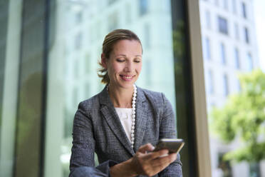 Lächelnde Geschäftsfrau, die ein Mobiltelefon in der Nähe einer Glaswand benutzt - PNEF02863