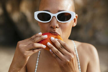 Woman wearing sunglasses eating mango at beach - EBSF03364