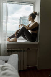 Nachdenkliche Frau sitzt zu Hause auf der Fensterbank - IEF00355