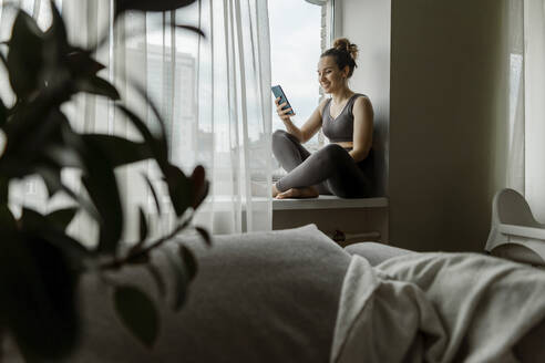 Frau schreibt Textnachrichten mit Telefon auf der Fensterbank zu Hause - IEF00358
