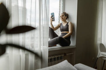 Woman using smart phone sitting on window sill at home - IEF00359