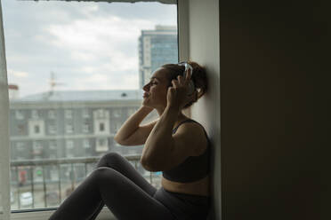 Frau genießt Musik mit Kopfhörern auf der Fensterbank zu Hause - IEF00360