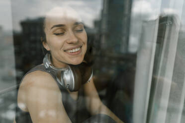 Happy woman with bluetooth headphones seen through glass window - IEF00364