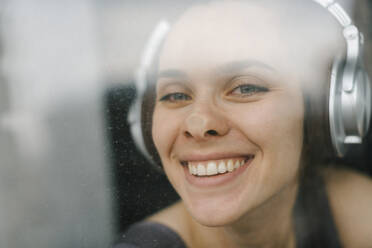 Lächelnde Frau mit Kopfhörern, die ihre Nase an ein Glasfenster drückt - IEF00384
