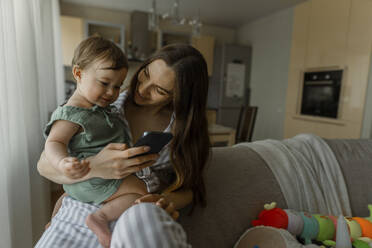 Mutter zeigt ihrem kleinen Mädchen im Wohnzimmer das Smartphone - IEF00398