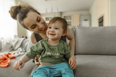 Mother dressing baby girl sitting on sofa in living room - IEF00407