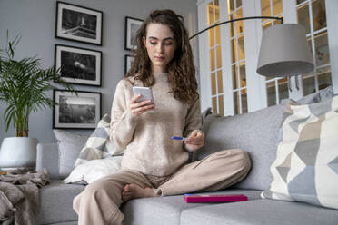 Young woman using smart phone holding pregnancy testing kit at home - VPIF08010