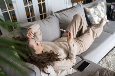 Young woman looking at pregnancy testing kit relaxing on sofa - VPIF08015