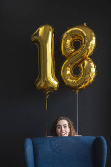 Surprised young woman hiding behind chair with number 18 balloon on black backdrop - VPIF08047