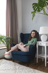 Smiling young woman relaxing on armchair at home - VPIF08055