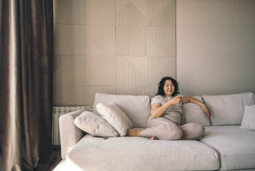 Woman holding glass of water sitting on sofa at home - ADF00072