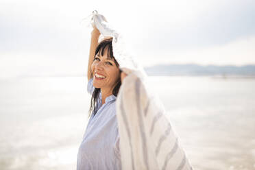 Cheerful woman holding scarf at beach - JOSEF19165