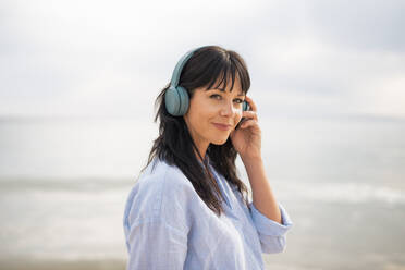 Mature woman with bangs wearing bluetooth headphones at beach - JOSEF19182