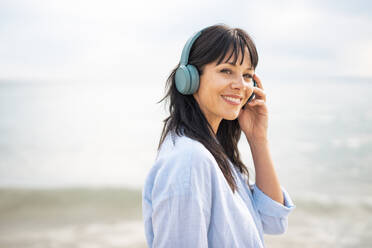 Lächelnde Frau mit drahtlosen Bluetooth-Kopfhörern am Strand - JOSEF19184