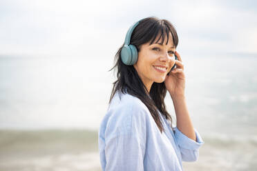 Smiling woman wearing wireless bluetooth headphones at beach - JOSEF19184