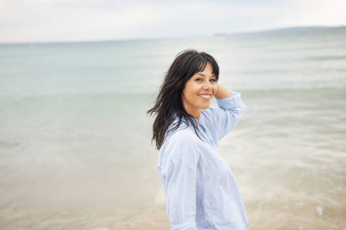 Smiling mature woman with hand in hair at beach - JOSEF19216