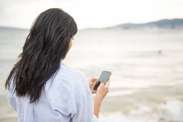 Frau mit langen Haaren benutzt Smartphone am Strand - JOSEF19220