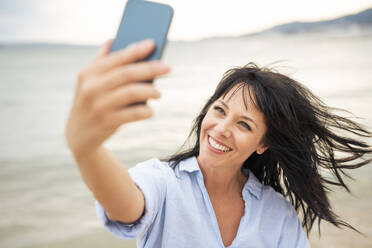 Eine fröhliche Frau mit zerzaustem Haar, die ein Selfie mit ihrem Smartphone macht, während sie den Strand genießt - JOSEF19224