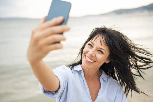 Happy woman with tousled hair taking selfie through smart phone at beach - JOSEF19224