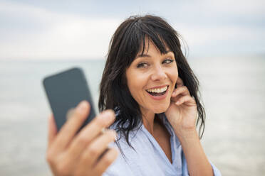 Eine glückliche Frau hält einen Moment mit ihrem Smartphone fest, während sie den Strand genießt - JOSEF19227